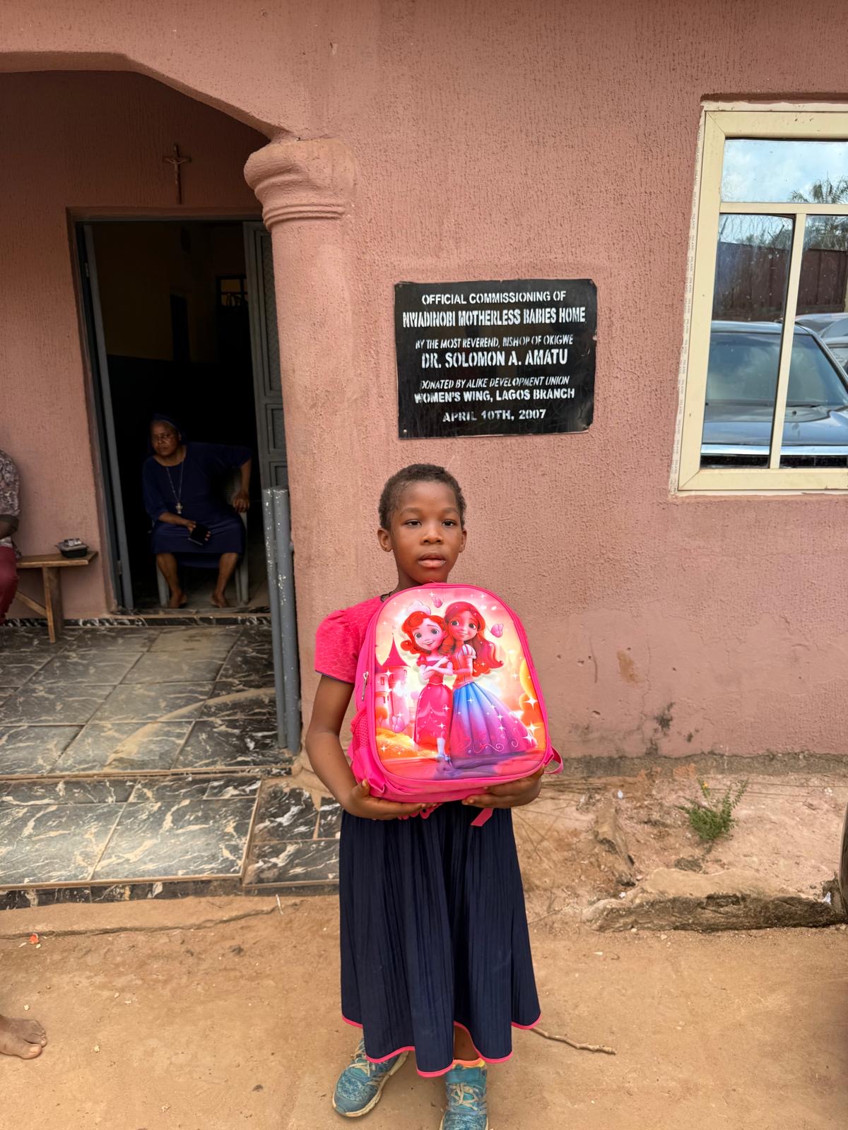 A child holding a pink backpack outside the orphanage during a support visit.