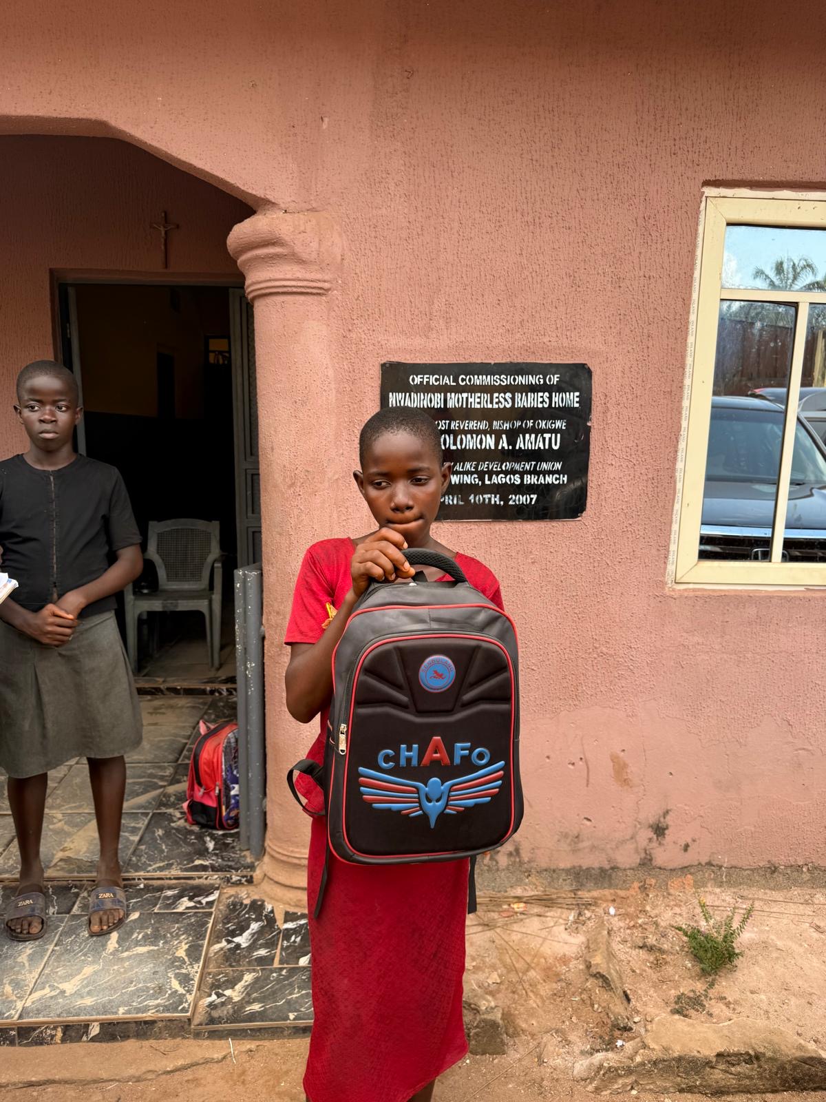 A child in red holding a backpack outside the orphanage.