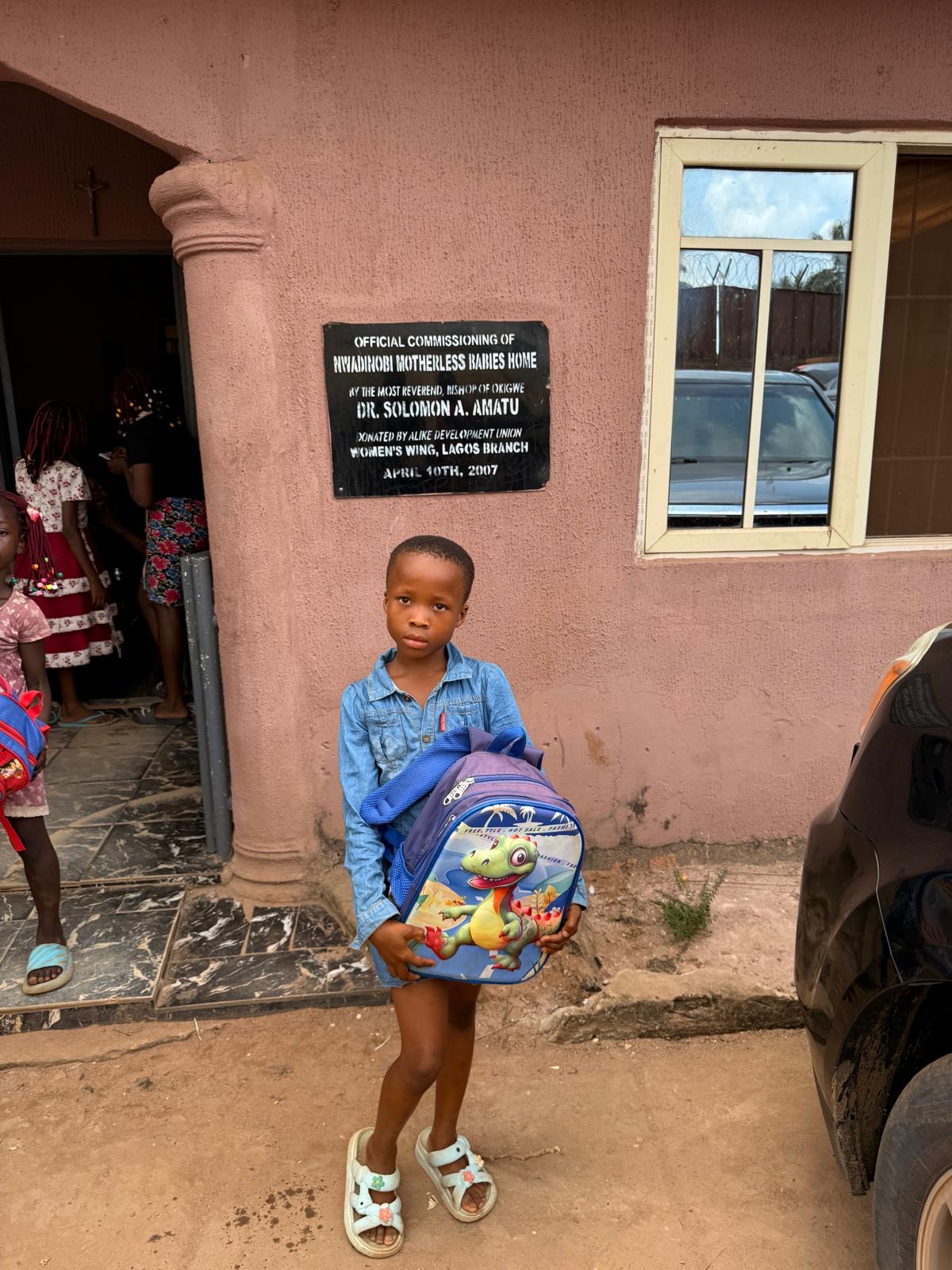 A young child holding a dinosaur backpack outside the orphanage.
