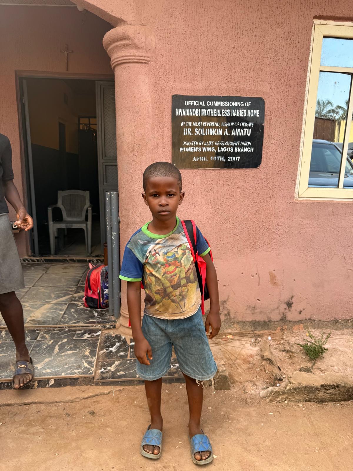 A young child standing outside the orphanage with a backpack.